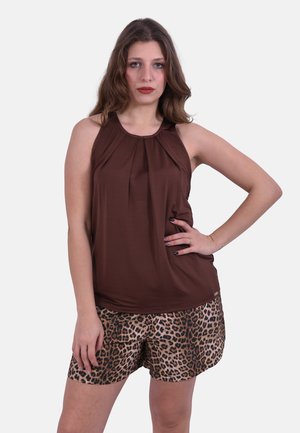 Donna con capelli castani lunghi che indossa una canotta marrone senza maniche e pantaloncini con stampa leopardata, in piedi con una mano sui fianchi.