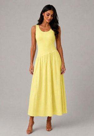 Femme aux longs cheveux foncés porte une robe maxi jaune sans manches avec un haut ajusté et une jupe fluide, debout devant un fond gris uni.