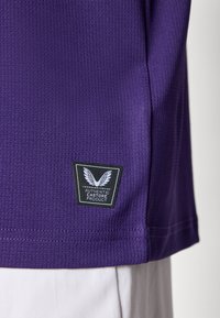 T-shirt de sport violet en tissu texturé, avec une étiquette noire. L'étiquette présente un logo blanc et un texte indiquant "PRODUIT AUTHENTIQUE CASTORE".