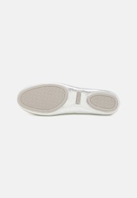 Suela de un zapato blanco plano con dos almohadillas de goma beige antideslizantes, una más grande en la parte delantera y una más pequeña en el talón, sobre un fondo blanco.