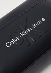 Crna imitacija kože s utisnutim logotipom "CK", srebrni tekst "Calvin Klein Jeans", glatka tekstura i šivani rubovi.