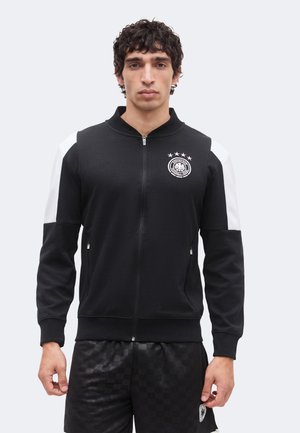 Mies, jolla on musta vetoketjullinen takki valkoisilla hartiapaneeleilla ja merkki, yhdistetty mustiin ruutukuviollisiin shortseihin yksiväristä taustaa vasten.