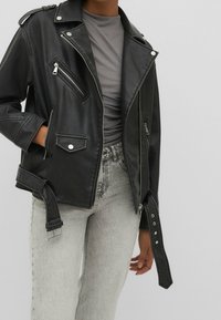 Veste de motard en simili cuir noir avec poches zippées, épaulettes et ceinture ajustable, associée à un jean gris clair et un haut gris.
