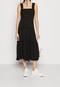Femme portant une robe midi noire sans manches avec jupe à volants, des baskets blanches et tenant un petit sac à bandoulière blanc.
