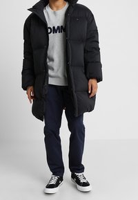 Giacca a piumino nera con collo alto, bottoni frontali e design trapuntato, indossata sopra un maglione grigio chiaro e pantaloni blu scuro, abbinata a sneaker.