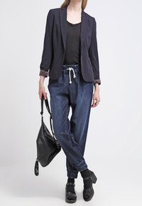 Tmavě modrý blejzr se vzorovanými manžetami, černé tričko, volné džíny v jogger stylu s elastickými manžetami a černé kotníkové boty, držící černou kabelku.