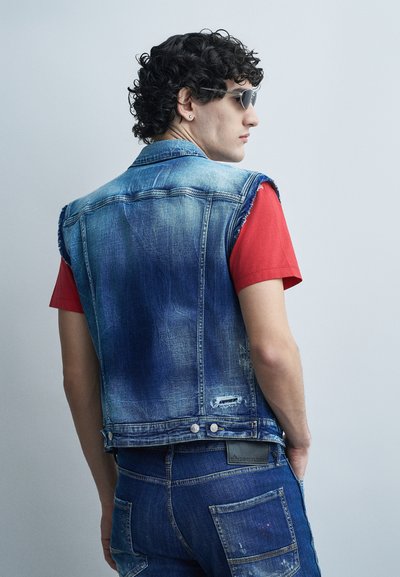 Jeune homme aux cheveux bouclés portant des lunettes de soleil noires, une veste en denim délavée sans manches sur un t-shirt rouge, et un jean en denim, de profil.