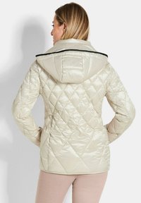 Giacca trapuntata beige con cappuccio, caratterizzata da cuciture a rombi e rifiniture nere lungo il colletto. Texture liscia e lucida.