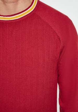 Pulover tricotat roșu cu un model texturat, având un guler rotund accentuat de dungi galbene și albe. Design cu mâneci raglan.