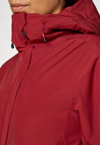 Rød vanntett jakke har høy krage med justerbare snorstrammere, glatt tekstur og frontlås med minimalistisk design.