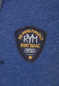 Tessuto blu navy con finitura texture, caratterizzato da una toppa a forma di scudo con "RYM" e "PORT ISAAC" ricamati in bianco e giallo.