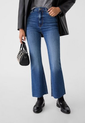 Femme portant un jean flare court bleu, mocassins en cuir noirs, veste noire, tenant un sac à main noir clouté sur un fond uni.