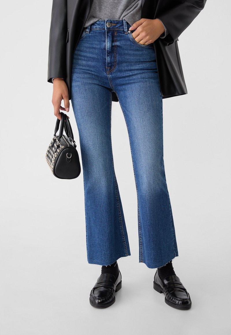 Femme portant un jean flare court bleu, mocassins en cuir noirs, veste noire, tenant un sac à main noir clouté sur un fond uni.