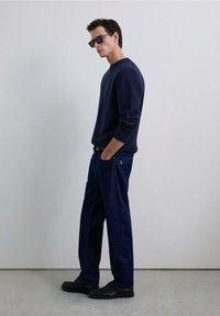 Jersey de punto navy, pantalones de mezclilla oscuros de corte relajado y zapatos negros. Los accesorios incluyen gafas de sol negras. Diseño simple, textura suave.
