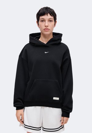 Person mit kurzen dunklen Haaren, die einen schwarzen Nike-Kapuzenpullover und weiße Shorts mit schwarzen Streifen trägt, steht vor einem schlichten Hintergrund.