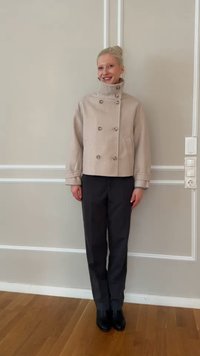 Χαμογελαστό άτομο με ξανθά μαλλιά, φορώντας μπεζ παλτό με κουμπιά, σκούρο παντελόνι και μαύρα παπούτσια, στέκεται μπροστά σε ανοιχτόχρωμο τοίχο μέσα σε κλειστό χώρο.