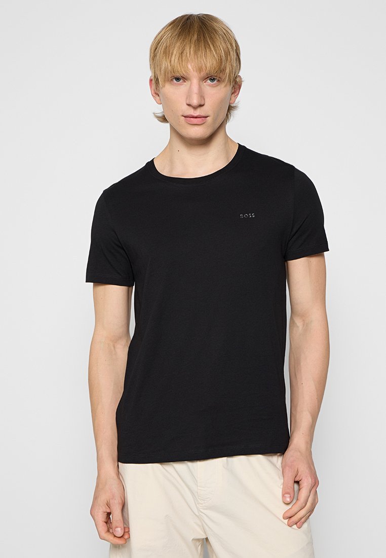 Junger Mann mit kurzem blonden Haar, der ein enges schwarzes BOSS-T-Shirt und beige Hosen trägt und vor einem schlichten weißen Hintergrund steht.