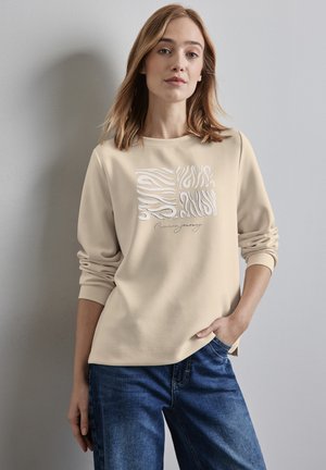 Sweatshirt beige avec motif abstrait blanc et détails en script, doté de manches longues et d'une coupe décontractée, associé à un jean en denim bleu.