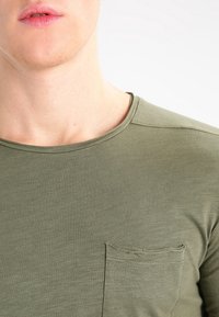 Camiseta de algodón verde oliva con cuello redondo, que presenta un pequeño bolsillo en el pecho y un acabado de tela texturizada.