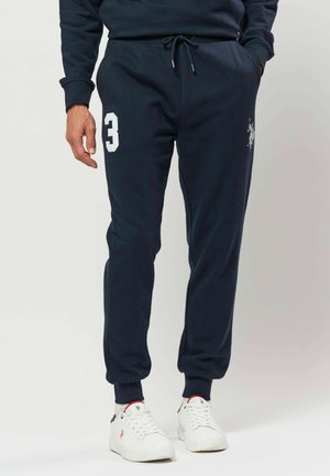 Homme portant un jogging bleu marine avec un numéro trois blanc et un logo, assorti à des baskets blanches avec des détails rouges et noirs.