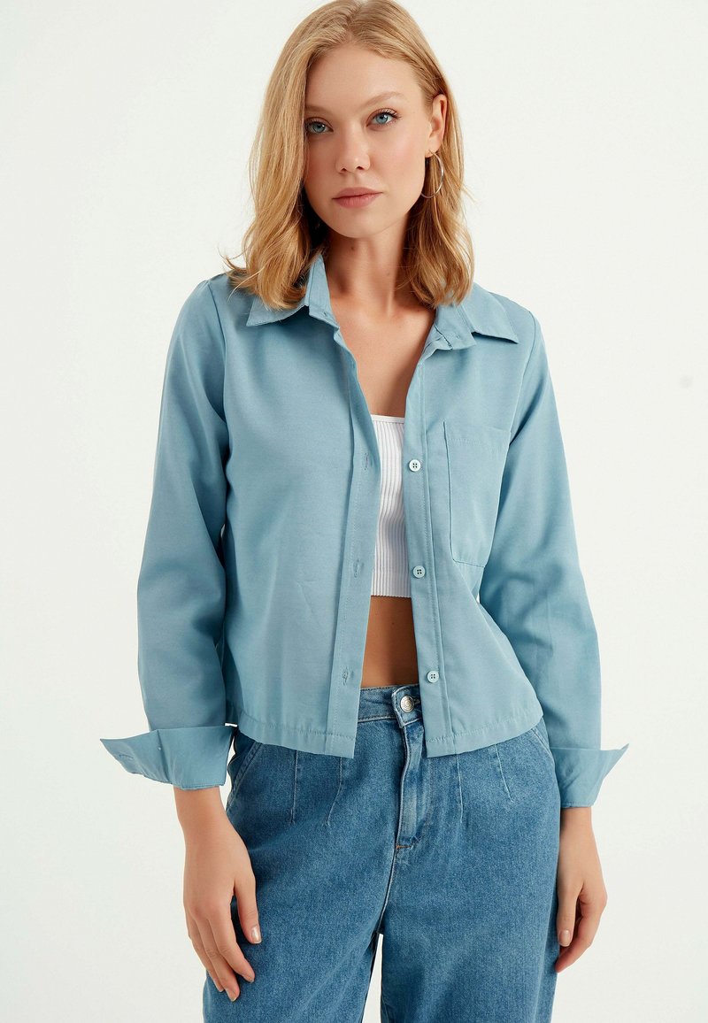 Hellblaues, kurz geschnittenes Hemd mit Knöpfen, Kragen, Brusttasche und hochgerollten Ärmeln, aus einem glatten Stoff. Kombiniert mit hochtaillierten Jeans.