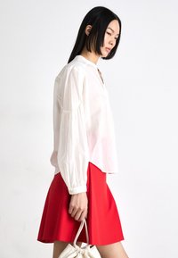 White long-sleeved blouse with a loose fit and buttoned cuffs, paired with a red flared skirt, held with a white handbag.