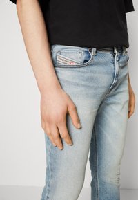 Bleka blå denimjeans med slim fit, med framfickor och en synlig varumärkesetikett på bakfickan. Handen vilar på låret.