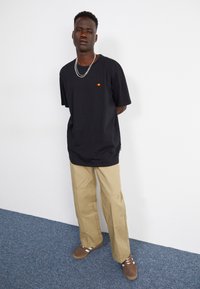 Schwarzes T-Shirt mit einem kleinen roten und gelben Logo, kombiniert mit weit geschnittenen, beigefarbenen Hosen. Die Schuhe sind braun mit weißen Streifen. Einfaches Design, Baumwollstoff.