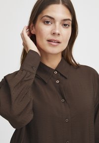 Brown button-up shirt with a soft, textured fabric, featuring a collar and long sleeves. Wooden buttons accent the front.