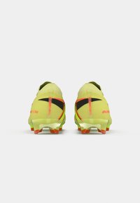 Jasne žlté futbalové kopačky s čiernymi a oranžovými akcentmi, ktoré majú aerodynamický dizajn, štruktúrovaný zvršok a priehľadnú podrážku s výraznými výstupkami.
