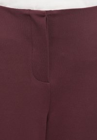 Pantalons bordeaux avec une texture lisse et un subtil détail de couture à l'arrière. Dotés d'une taille ajustée et sans matériel ou embellissements visibles.