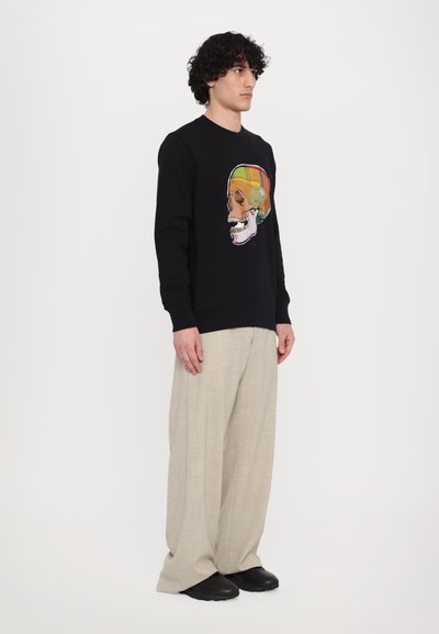 Sort sweatshirt med en farverig kraniumillustration på forsiden, parret med lyse beige brede bukser og sorte sko.