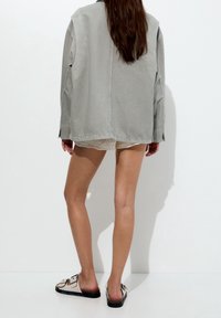 Vaaleanharmaa ylisuuri takki, jossa on pitkät hihat, teksturoitu kangas ja sivuhiat. Yhdistetty beigeihin pitsishortseihin ja mustiin sandaaleihin.