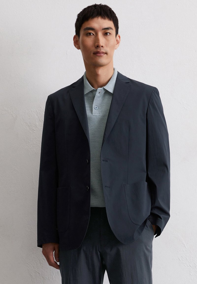 Granatowa marynarka z wyraźnymi klapami i dwiema przednimi kieszeniami, noszona na jasnoniebieskiej koszulce polo z kołnierzykiem, wykonanej z tkaniny o teksturowanej powierzchni.