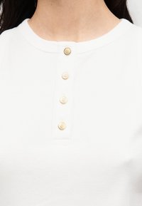 Λευκό βαμβακερό τοπ με στρογγυλή λαιμόκοψη και τέσσερα χρυσά κουμπιά κατά μήκος του παλκέ, με απαλή υφή και εφαρμοστή σχεδίαση.