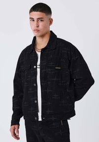 Chaqueta vaquera negra con un patrón sutil y cierre de botones, con bolsillos en el pecho y un corte relajado. Llevada sobre una camiseta blanca.