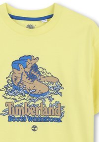 Geel T-shirt met een grafisch ontwerp van bruine laarzen met blauwe accenten en de tekst "Timberland Boots With Roots" in vetgedrukte, stijlvol lettertype.