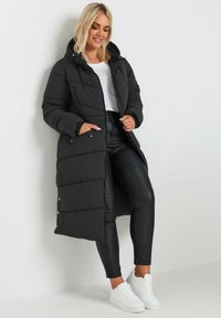 Zwarte lange pufferjas met capuchon, voorzien van een gewatteerd ontwerp, zijzakken en drukknoopsluiting. Gemaakt boven een zwarte leren broek en witte sneakers.