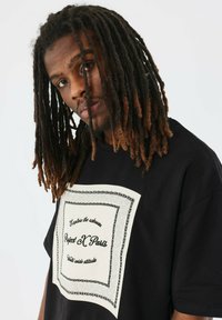 Camiseta de algodón negra con una gran impresión gráfica blanca que presenta detalles de borde intrincados en negro y texto en una mezcla de fuentes.