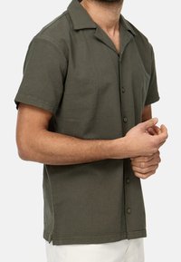 Camisa de manga corta color verde oliva con frente abotonado, cuello extendido y aberturas laterales para mayor comodidad de movimiento. Material de algodón con un acabado texturizado.