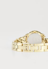 Gold metal watch featuring a polished link bracelet, a round case, and a silver-tone dial. Brand name “GUESS” engraved on links.