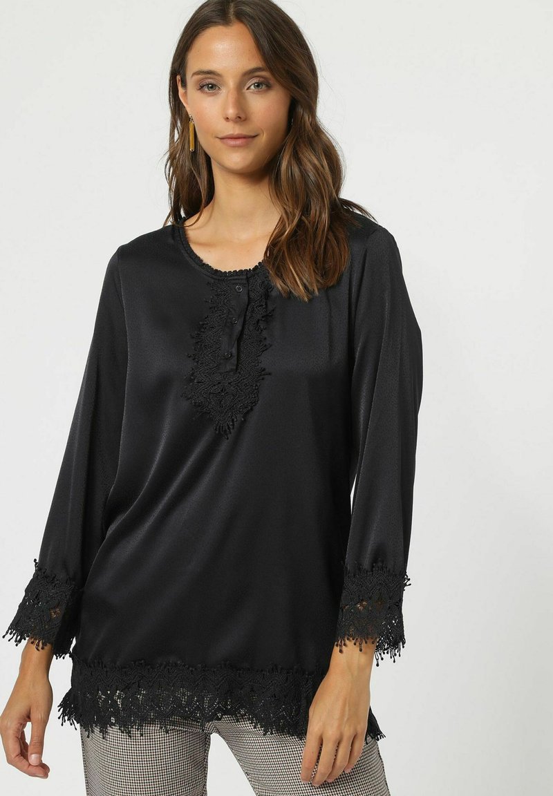 Blusa de satén negra con mangas largas, un borde de encaje en el escote y los puños, y una serie de botones en la parte delantera. Textura suave.