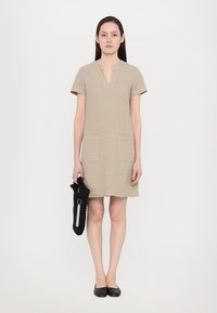 Korte mouwen linnen jurk in beige met een V-hals. Beschikt over twee voorzakken en een ontspannen pasvorm. Model houdt een zwarte textuur tas vast.