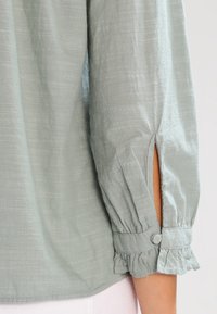 Light green blouse with long sleeves, gathered cuffs, and side slits. Soft texture with a slight sheen and button accents on the cuffs.