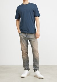 Námořnické tričko s kulatým výstřihem a krátkými rukávy, s nenápadným logem. Pár je se světle modrými, vybledlými džíny a bílými teniskami.