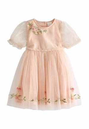 Bambino in un vestito pesca con ricami floreali si trova su un'altalena di legno incorniciata da foglie verdi e fiori in un giardino.