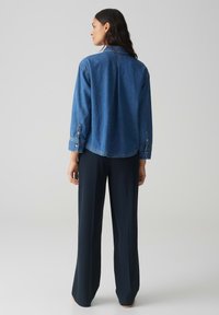 Giacca di denim blu medio con maniche lunghe, polsini abbottonati e vestibilità rilassata. Indossata sopra pantaloni larghi blu navy, che mostrano uno stile casual.