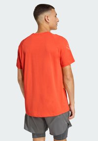 Camiseta deportiva roja de manga corta, con cuello redondo y sutiles detalles en los hombros, combinada con pantalones cortos deportivos grises.