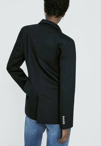 Blazer noir à coupe ajustée, col à revers cranté, fente arrière et détails de boutons sur la manche, associé à un jean en denim bleu clair.