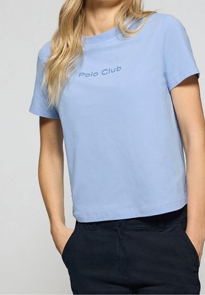 Kobieta w jasnoniebieskiej koszulce z krótkim rękawem z napisem "Polo Club" i ciemnych spodniach, z rękami w kieszeniach, na neutralnym tle.